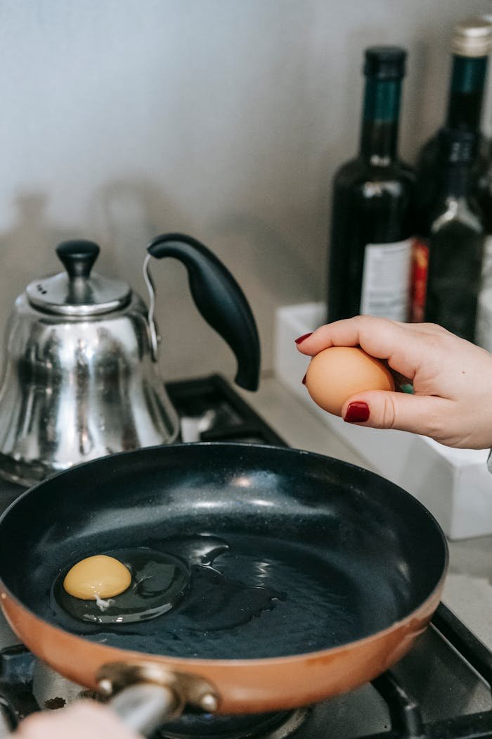 A person's hand cracking an egg into a frying pan on a stovetop with kitchen items.